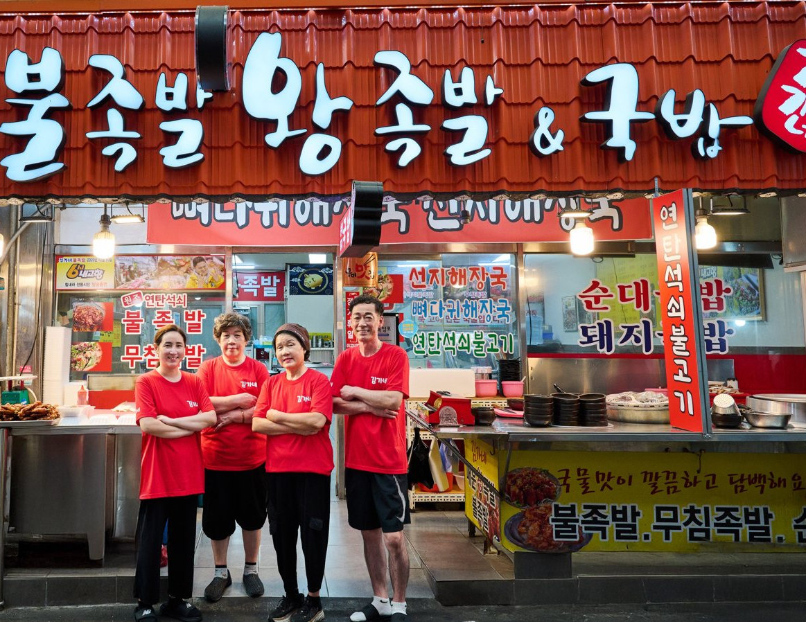 시장 가게 앞에 빨간 '김가네' 티셔츠를 입은 네 사람이 팔짱을 끼고 서 있다 — 위로 '김가네 불족발 왕족발 & 국밥' 간판, 뒤로 차림판·족발 쟁반