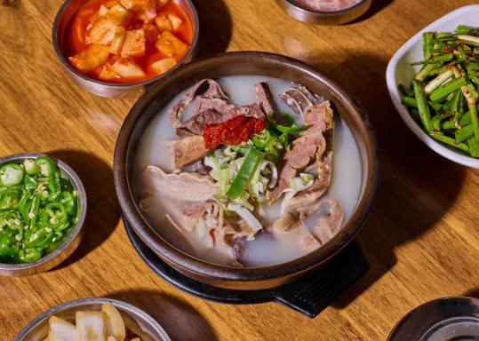 검은 뚝배기의 뽀얀 국물 돼지국밥 — 돼지고기·내장 건더기, 붉은 다대기, 파·부추. 둘레에 깍두기, 썬 청양고추, 부추무침, 양파
