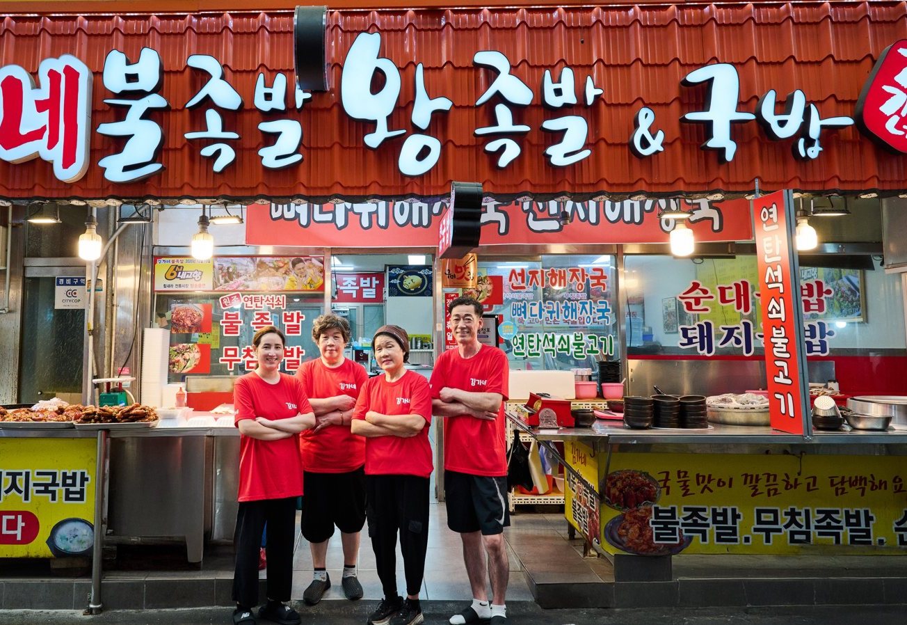 시장 가게 앞에 빨간 티셔츠를 입은 네 사람이 팔짱을 끼고 서 있다 — 위로 가게 간판, 뒤로 족발 쟁반